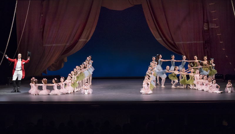 Child dancers in pastel colored tutus pose in cascading linear formations with their hands on one another's shoulders. Ask la Cour in a red tail coat and top hat stands off to the side of them.